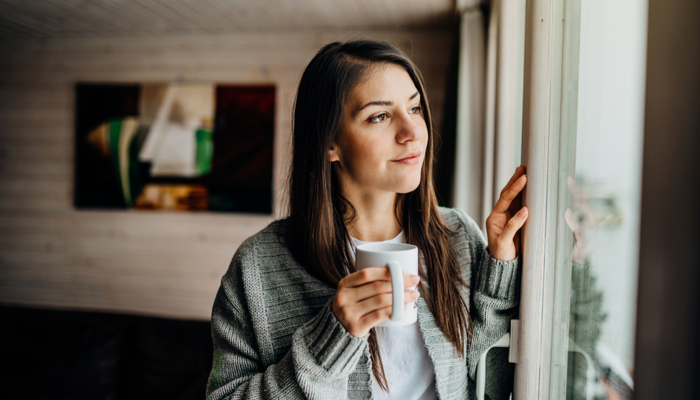Young woman ina cardigan with mug in hand spending free time home, staying home, enjoying view,gazing through to the window while remembering the good things