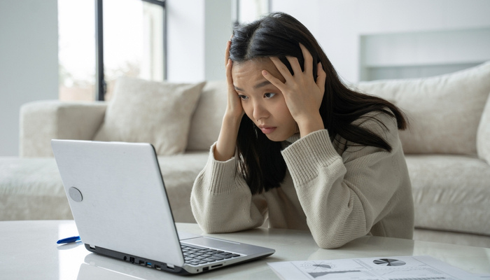 Exhausted Asian woman in sweatshirt touching her head with a gray laptop in the home office in the living room
