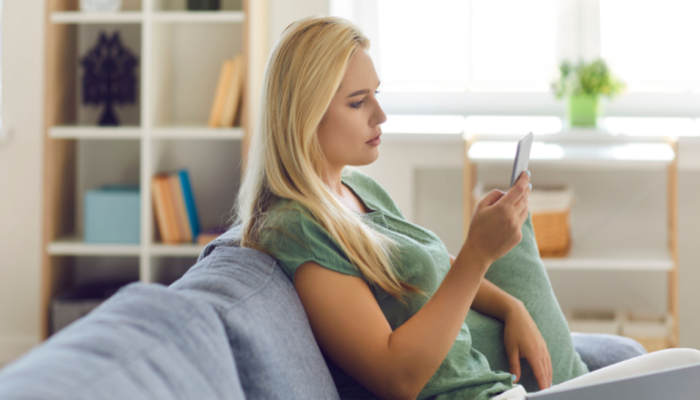 Side view of blonde woman sitting on a gray sofa at home, using cellphone, chatting on social media, reading texts, shopping in online store