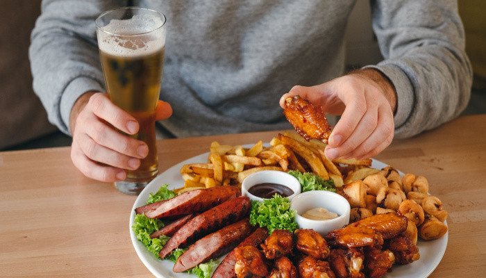 Man having dinner holding chicken leg and beer, large plate with sausages, fries, chicken and sauses on the table
