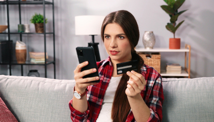 Happy cheerful, smiling brunette woman dreaming of new dress, looking at the smartphone with rejoice while holding credit card and making purchase